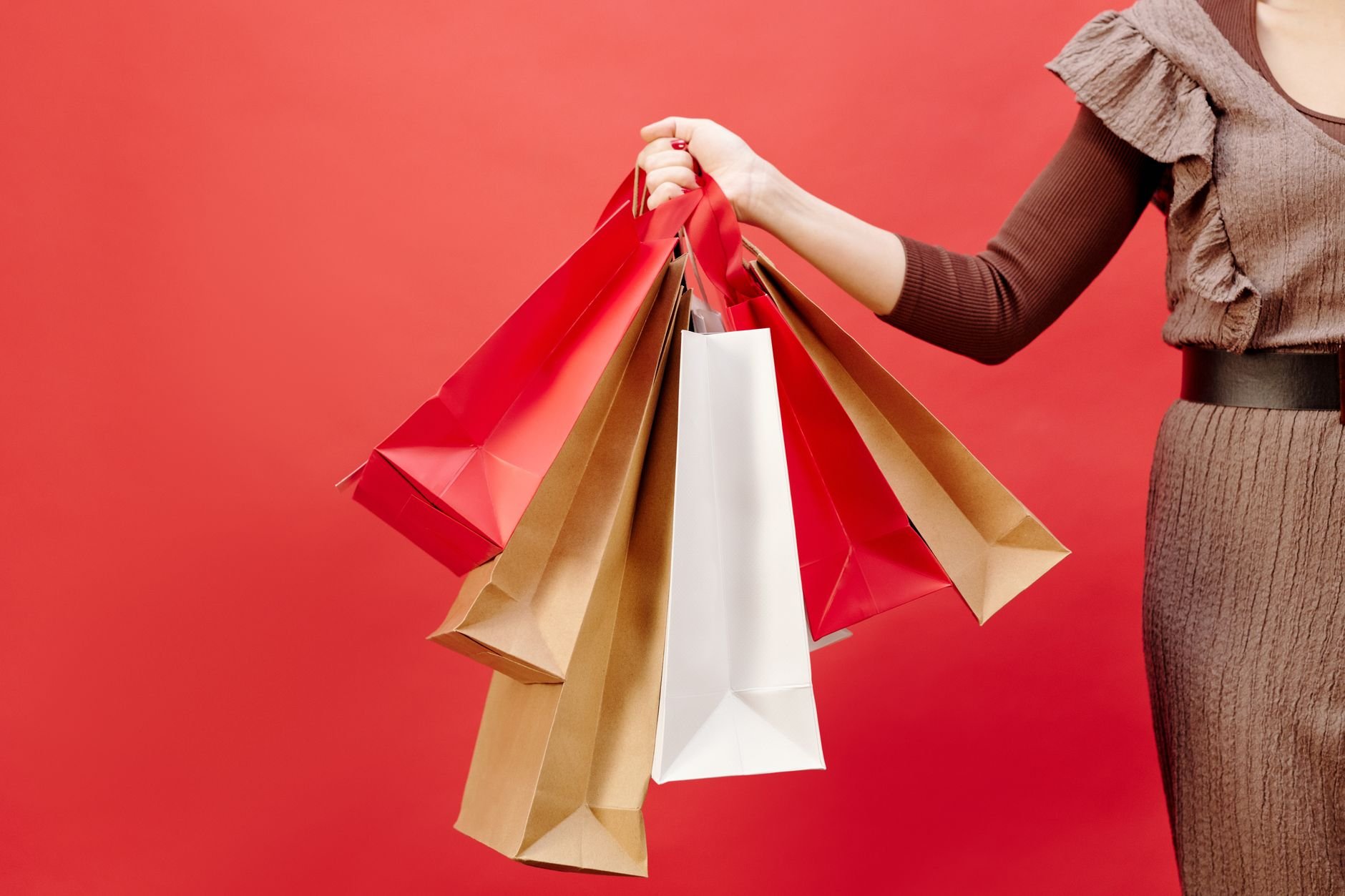 Mulher de vestido marrom segurando várias sacolas de compras diante de um fundo vermelho vivo.